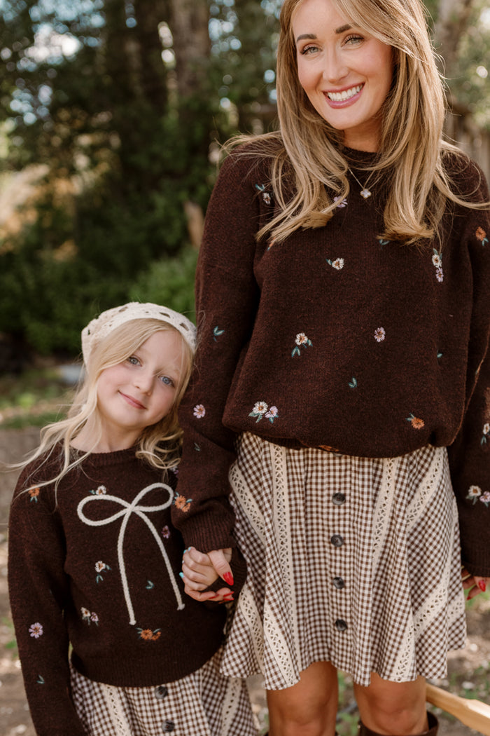 A smiling woman and young girl hold hands, both wearing matching Ivy City Co Mini Ellis Sweaters in Warm Cocoa Lovelorn Ditsy with charming embroidered details and balloon sleeves, paired with plaid skirts as they stand outdoors among trees.