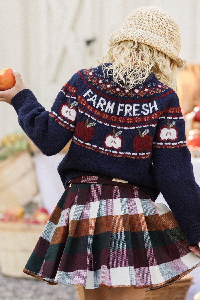 A person with curly blond hair wears a straw hat and the Ivy City Co Mini Farm Fresh Sweater, featuring playful apple designs and stylish balloon sleeves, paired with a plaid skirt while holding an apple. The backdrop is filled with baskets of produce, evoking the charm of an outdoor market or farm setting.