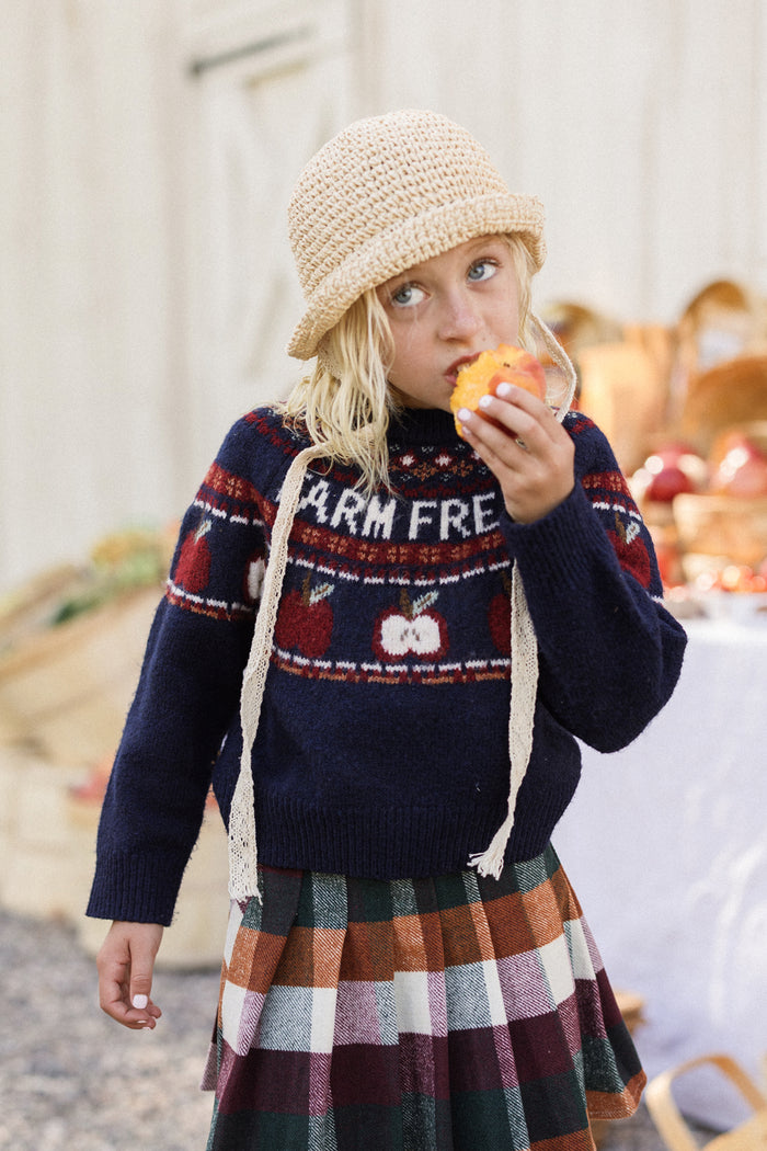 A child in a straw hat and the Ivy City Co Mini Farm Fresh Sweater enjoys an apple outdoors, surrounded by wooden crates and a table of apples in the background.