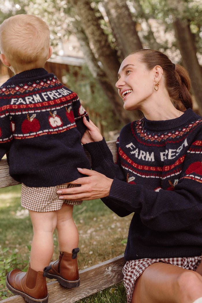 A woman and a small child sit outdoors on a wooden fence, smiling and enjoying a sunny day among trees. The child is wearing the Ivy City Co Mini Farm Fresh Sweater, featuring adorable balloon sleeves that add extra charm to their coordinated look.