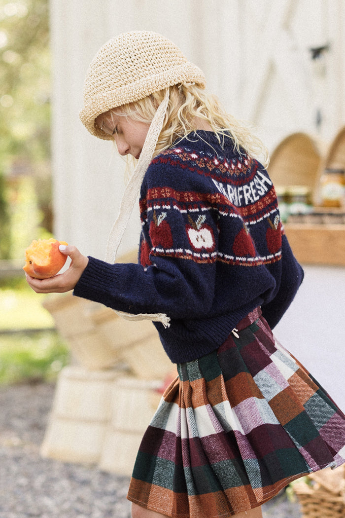 A child wearing the Ivy City Co Mini Farm Fresh Sweater and a plaid skirt holds a peach while walking outdoors near wooden crates and a rustic stand.