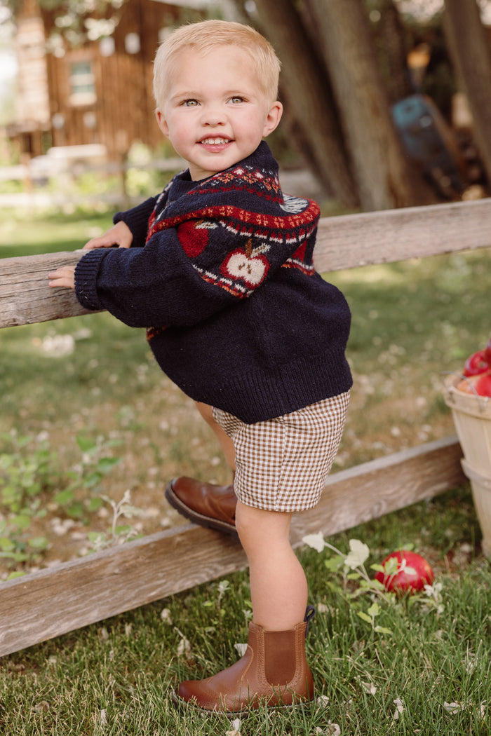 A smiling toddler stands on the grass near a wooden fence, one foot raised, wearing the Ivy City Co Mini Farm Fresh Sweater—an adorable navy sweater dress featuring playful animal motifs and charming balloon sleeves—paired with plaid shorts and brown boots. A nearby basket filled with apples completes the cheerful outdoor scene.