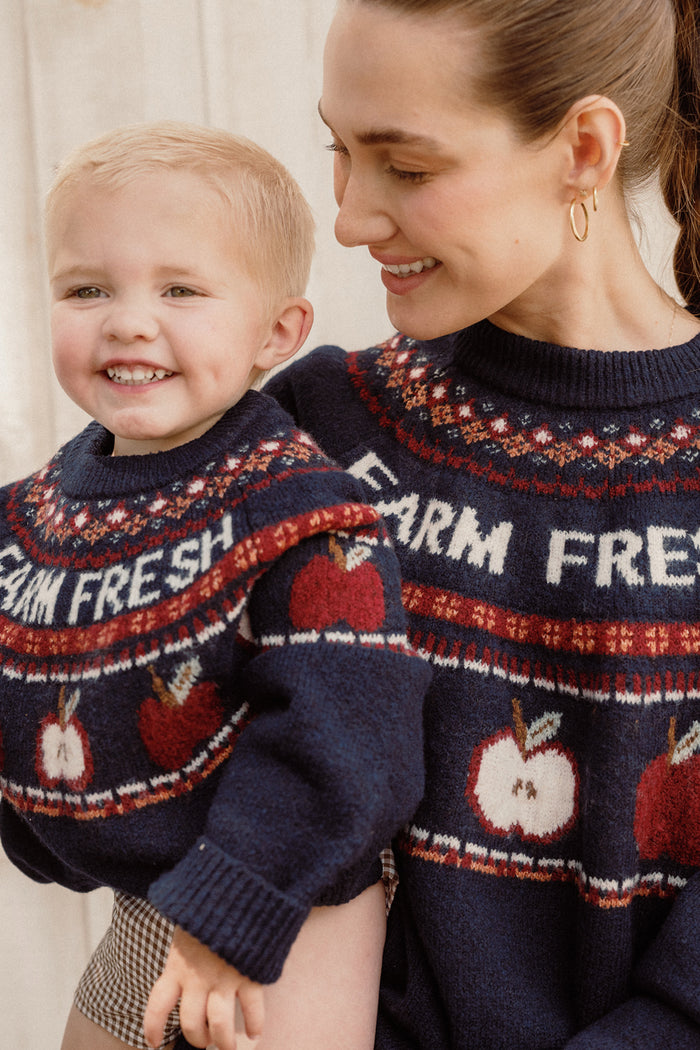 A smiling woman and child wear matching navy Ivy City Co sweaters featuring playful apple designs and the words "FARM FRESH." The woman embraces the laughing child, whose Mini Farm Fresh Sweater dress stands out with its charming balloon sleeves, completing their adorable coordinated look.