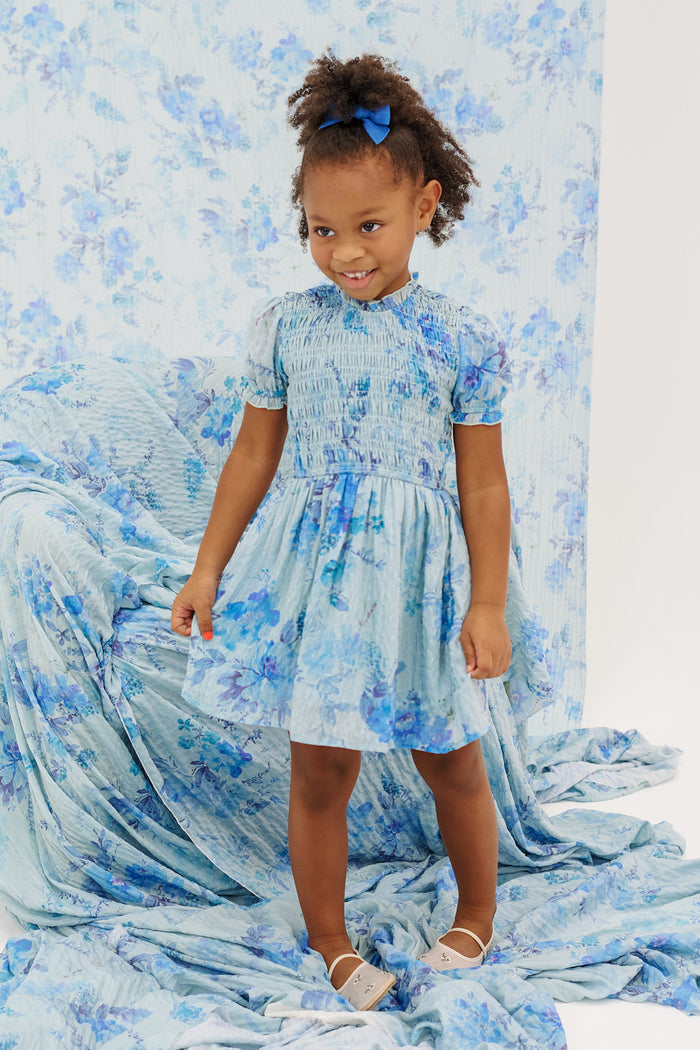 A young girl smiles in front of a blue floral backdrop, wearing the Ivy City Co Mini Flynn Dress in Aquarelle Fleur with a bow in her hair and white sandals—perfect for spring or mommy and me matching looks.