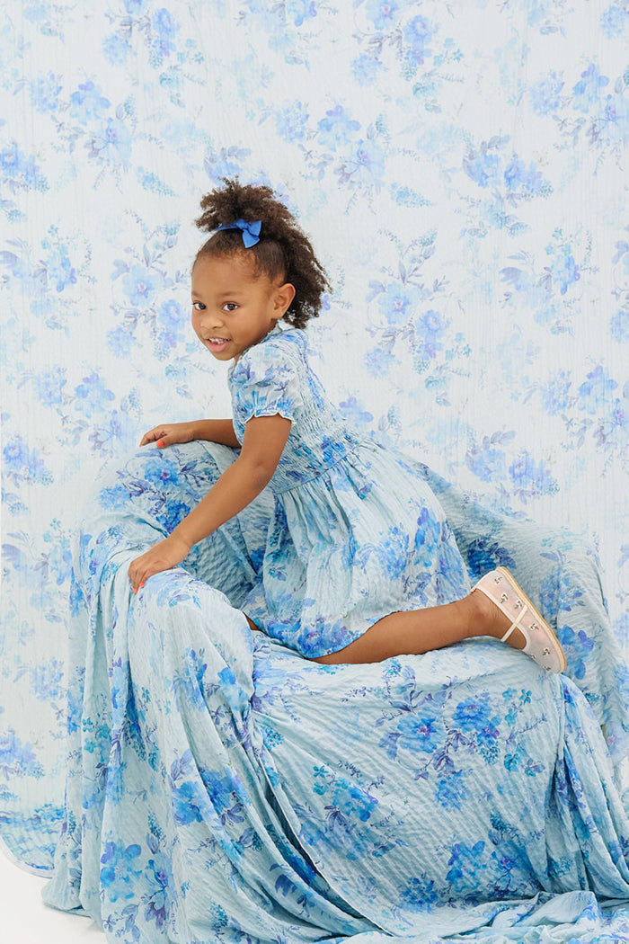 A young girl in Ivy City Co’s Mini Flynn Dress in Aquarelle Fleur with a matching hair bow kneels on a floral-draped chair, blending into the background—capturing the perfect spring look for girls.