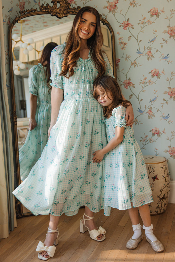 A woman and a young girl, both in matching Ivy City Co Mini Jessie Dresses in Bloom Gingham, smile together by an ornate mirror in a floral-wallpapered room as the girl hugs the woman's waist.