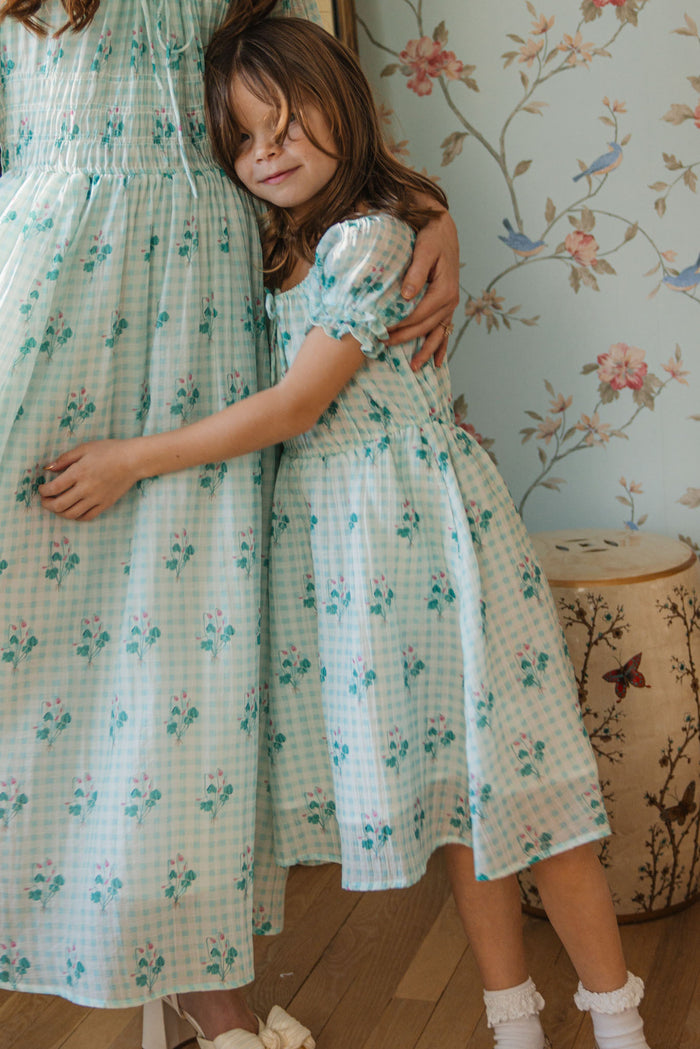 A young girl in the Mini Jessie Dress in Bloom Gingham by Ivy City Co hugs an adult in a matching dress, standing before floral wallpaper and a decorative stool. The girl’s eyes are closed, and she looks content.
