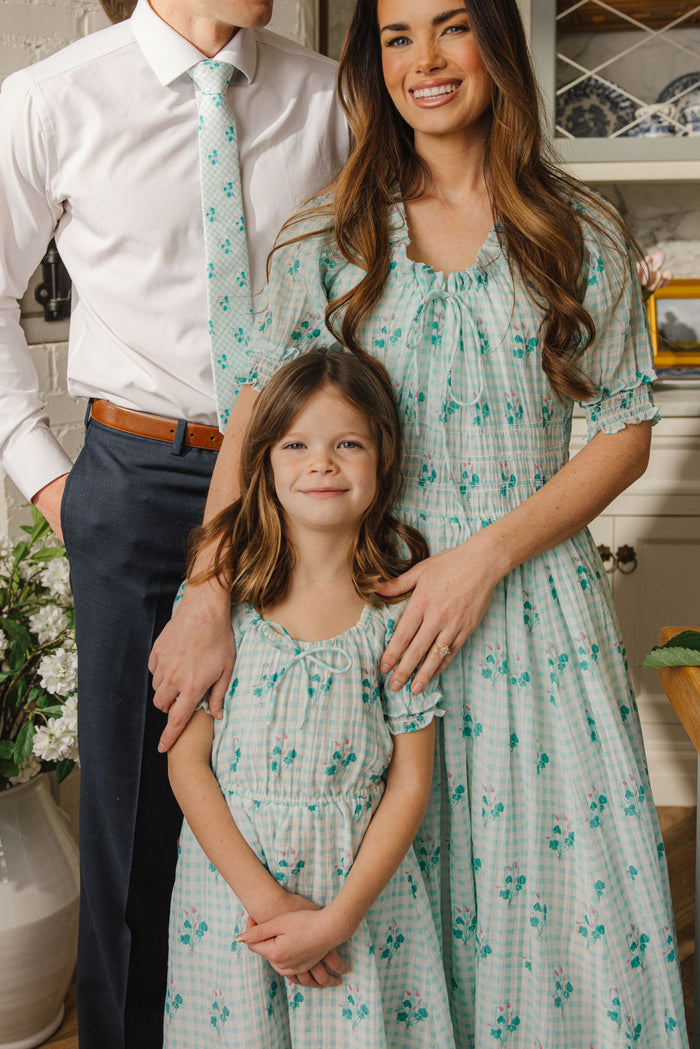 A man, woman, and young girl stand together indoors. The woman and girl wear matching Ivy City Co Mini Jessie Dresses in Bloom Gingham, while the man pairs a white shirt and blue trousers with a floral tie. All appear happy.