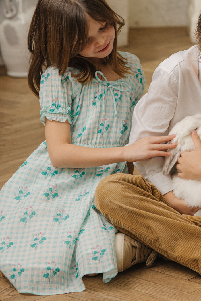 A young girl wears the Ivy City Co Mini Jessie Dress in Bloom Gingham as she gently pets a white rabbit on the lap of someone in brown corduroy pants, both seated indoors on a wooden floor.