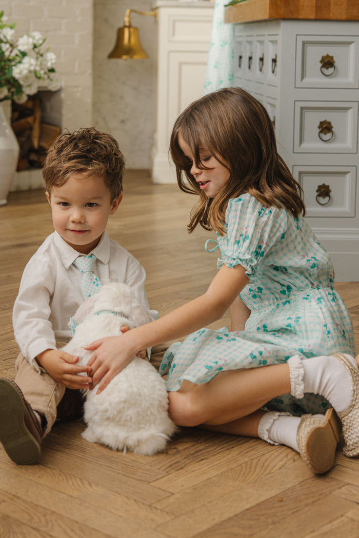 A young girl wears the Mini Jessie Dress in Bloom Gingham by Ivy City Co as she and a boy pet a white rabbit on a wooden floor, with a kitchen visible in the background.