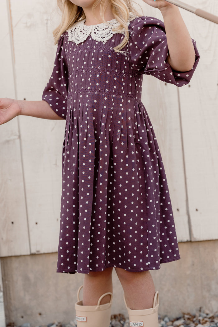 A young child stands outdoors holding a stick, dressed in the Ivy City Co Mini Jillian Dress in Aubergine Ditsy Dot, which features a white lace collar and charming puff sleeves. The child's face is not visible, and they are wearing beige rain boots.