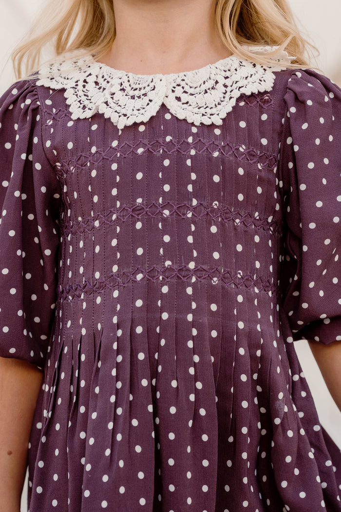 A child wearing the Ivy City Co Mini Jillian Dress in Aubergine Ditsy Dot, featuring a delicate white lace collar. The upper half of the viscose dress and neckline are visible, highlighting its subtle puff sleeves and charming purple fabric with white ditsy dots.