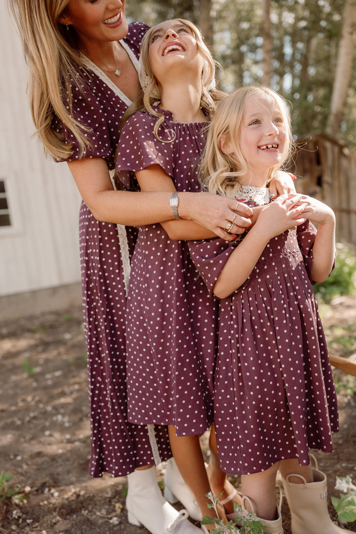 A woman and two young girls, all dressed in matching Ivy City Co Mini Jillian Dresses in Aubergine Ditsy Dot with charming puff sleeves and boots, stand outside smiling and laughing together in a sunny, wooded area.