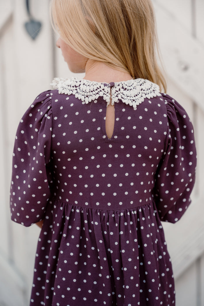 A girl with blonde hair stands with her back to the camera, wearing the Ivy City Co Mini Jillian Dress in Aubergine Ditsy Dot, featuring puff sleeves and a lace collar.