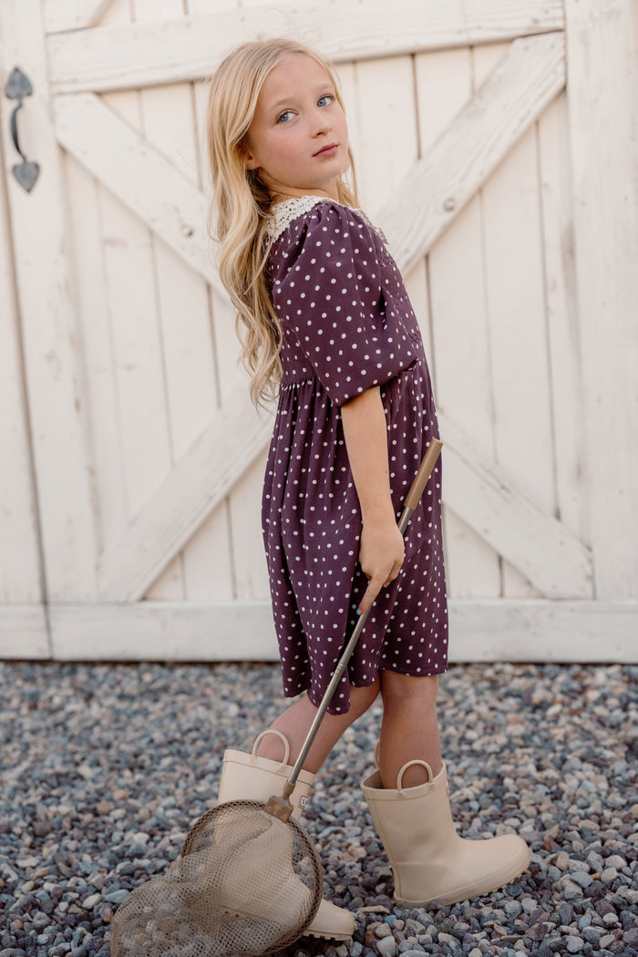 A young girl with long blonde hair wears the Ivy City Co Mini Jillian Dress in Aubergine Ditsy Dot, featuring viscose puff sleeves, and beige boots as she stands on gravel holding a net in front of a white wooden barn door.