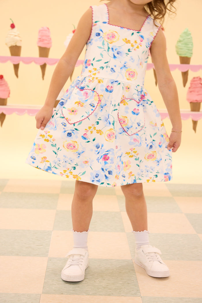 A young girl in the Ivy City Co Mini Rue Play Dress in Blue Bouquet with heart-shaped pockets and white shoes stands before a wall decorated with colorful ice cream cone cutouts.