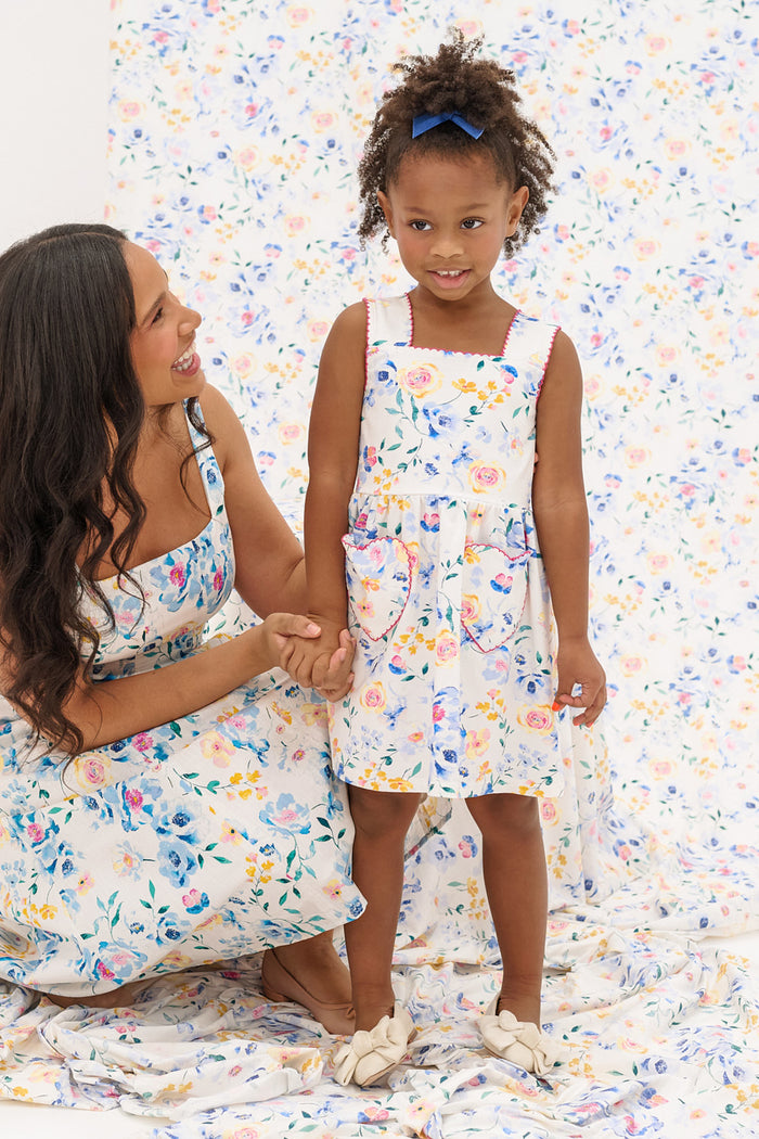 A woman kneels beside a young girl, both wearing Ivy City Co's Mini Rue Play Dress in Blue Bouquet, featuring pastel floral prints. The smiling woman and proud girl with a blue hair bow pose against a vibrant floral backdrop.
