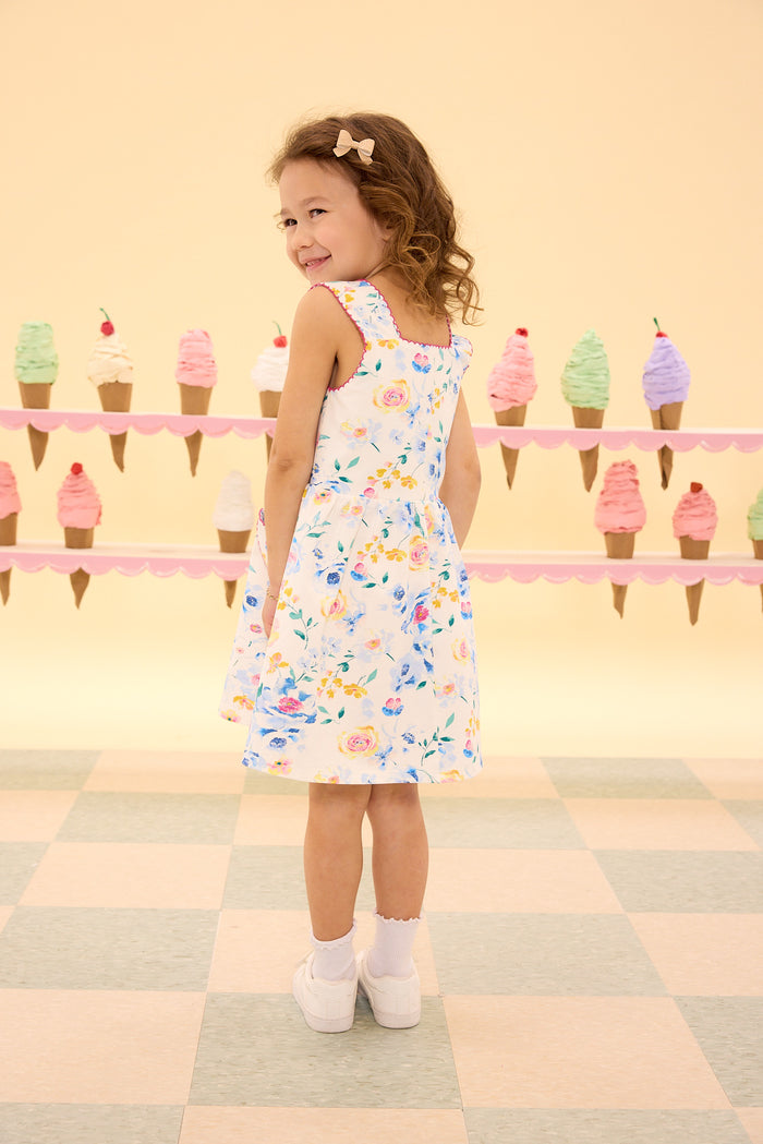 A young girl wearing the Ivy City Co Mini Rue Play Dress in Blue Bouquet and white shoes smiles over her shoulder. Behind her, colorful ice cream cone decorations sit on pastel pink shelves against a pale yellow wall.