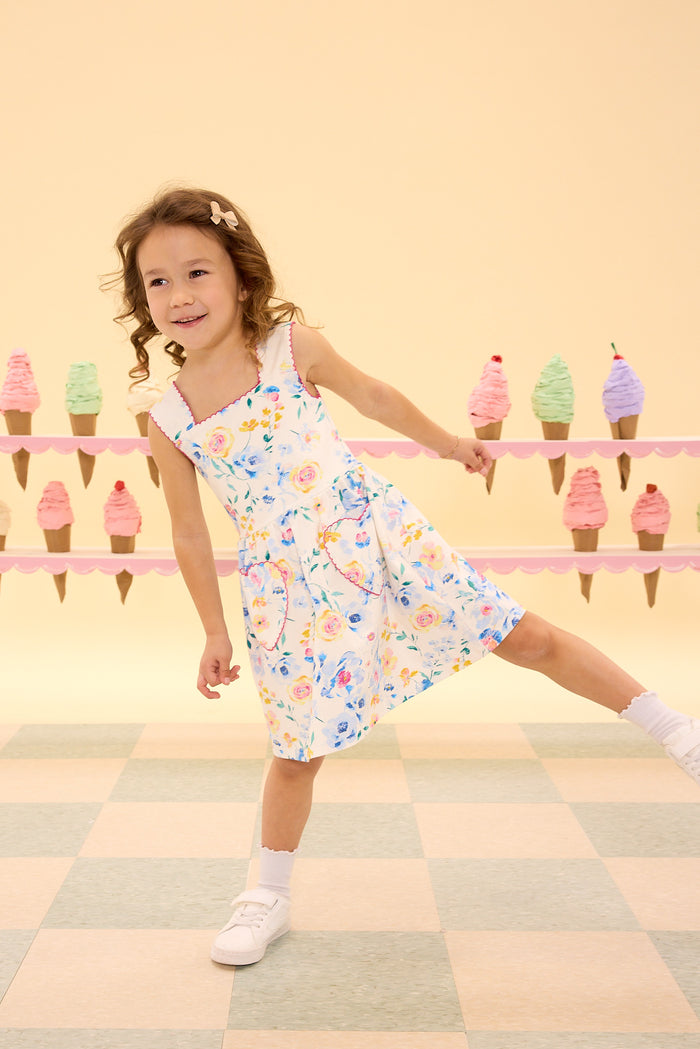 A young girl wears the Ivy City Co Mini Rue Play Dress in Blue Bouquet and white sneakers, standing playfully on one foot before a pastel backdrop adorned with colorful ice cream cone illustrations.