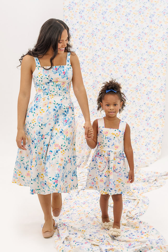 A woman and a young girl smile while holding hands in matching Ivy City Co Mini Rue Play Dresses in Blue Bouquet, standing together on coordinating floral fabric with a white background.