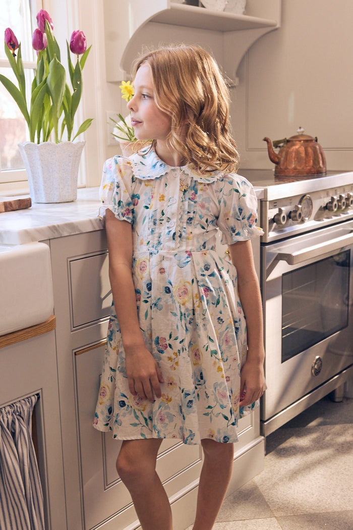 A young girl in the Ivy City Co Mini Sophia Dress in Blue Bouquet stands by a sunlit kitchen counter, gazing out the window. Fresh tulips, a teapot on the stove, and soft light enhance the tranquil scene.