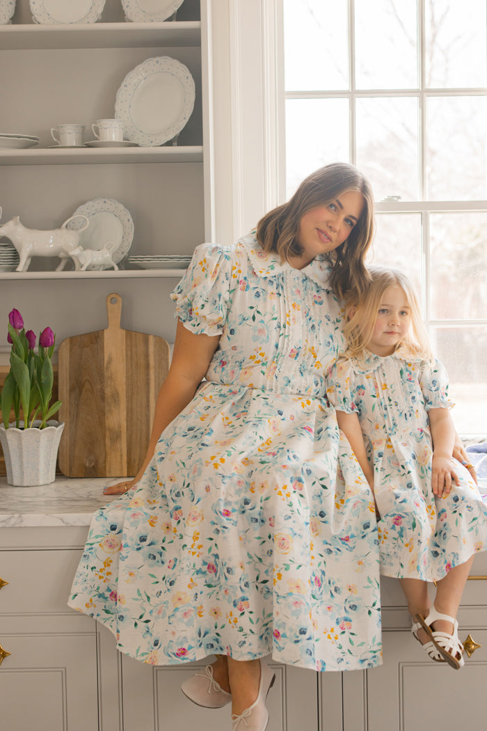 A woman and a young girl, both in the Mini Sophia Dress in Blue Bouquet by Ivy City Co, sit together on a kitchen counter. Plates line the shelves behind them and a pot of purple tulips rests nearby in the bright, cozy kitchen.