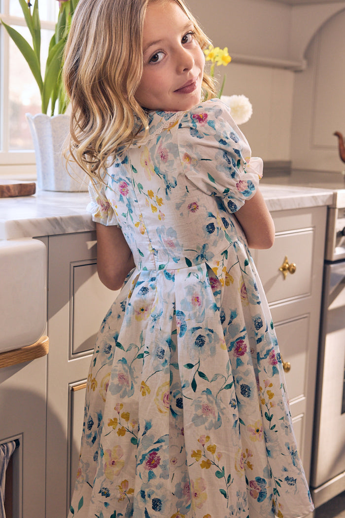 A young girl with wavy blonde hair wears the Mini Sophia Dress in Blue Bouquet by Ivy City Co, standing in a bright kitchen with sunlight streaming through the window and flowers on the counter. She looks back over her shoulder, smiling softly.