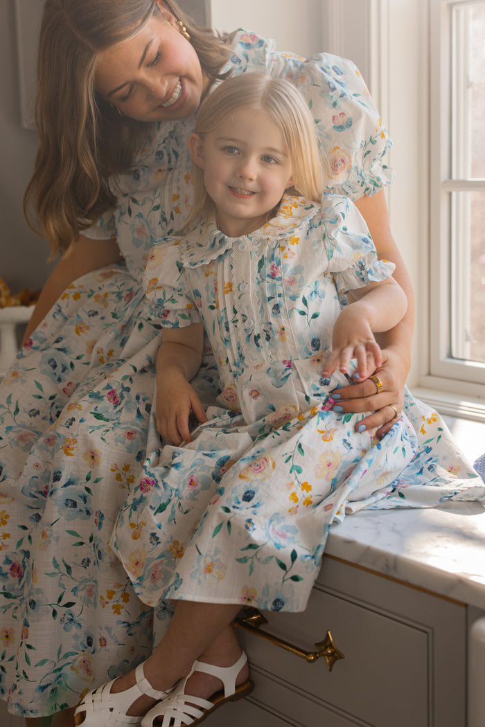 A woman and a young girl, both in Ivy City Co’s Mini Sophia Dress in Blue Bouquet, sit on a windowsill. The woman smiles at the girl, who grins at the camera as sunlight pours through the window.