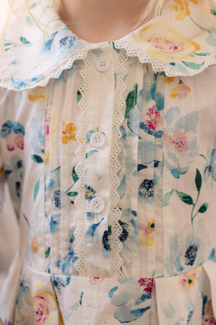 Close-up of a child wearing the Ivy City Co Mini Sophia Dress in Blue Bouquet, featuring pastel blue, yellow, and pink floral print, white buttons, lace trim, and a large rounded collar; only the lower face and chest are shown.