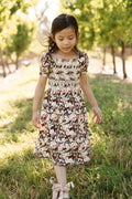 A young girl wearing the Ivy City Co Mini Sunday Best Dress in Midnight Garden, featuring a smocked bodice and puff sleeves, walks on grass in a sunlit outdoor setting with green trees in the background.