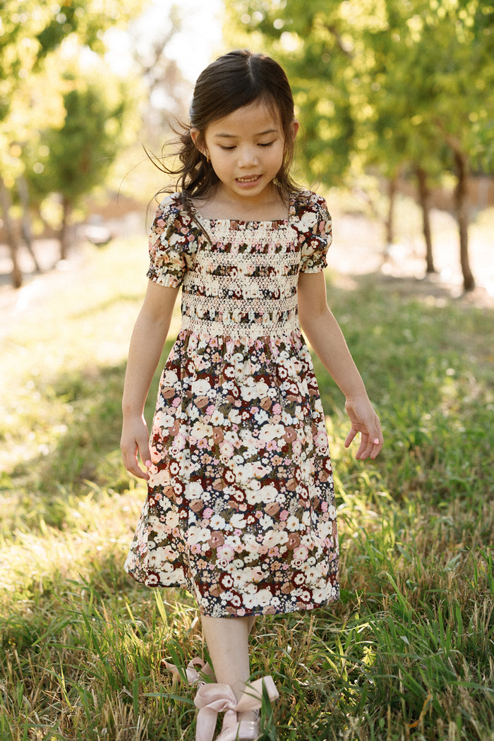 A young girl wearing the Ivy City Co Mini Sunday Best Dress in Midnight Garden, featuring a smocked bodice and puff sleeves, walks outdoors on green grass amid trees and sunlight, looking down with a gentle expression.