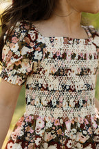 A child wearing the Ivy City Co Mini Sunday Best Dress in Midnight Garden, featuring puff sleeves, a floral pattern, and cream lace smocking on the bodice, stands outdoors with only the upper body and part of the face visible.