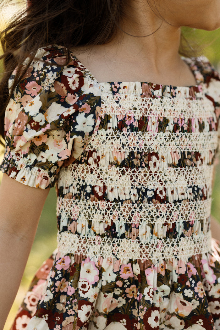 A child wearing the Ivy City Co Mini Sunday Best Dress in Midnight Garden, featuring puff sleeves, a floral pattern, and cream lace smocking on the bodice, stands outdoors with only the upper body and part of the face visible.