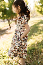A young girl wearing the Ivy City Co Mini Sunday Best Dress in Midnight Garden, featuring puff sleeves, stands outdoors on the grass with sunlight illuminating her as she looks down, hands tucked into her pockets. Trees and greenery create a lush backdrop behind her.