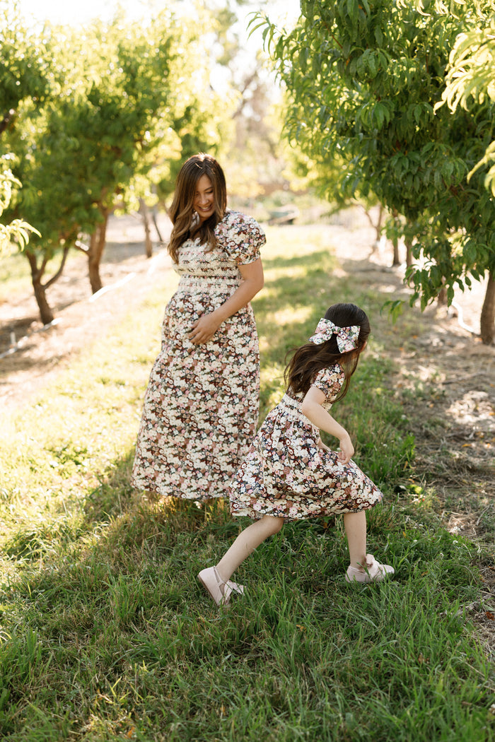 A woman and a young girl, both in Ivy City Co’s Mini Sunday Best Dress in Midnight Garden—featuring charming puff sleeves and a smocked bodice—are walking and playing together in a sunny orchard surrounded by lush green trees.