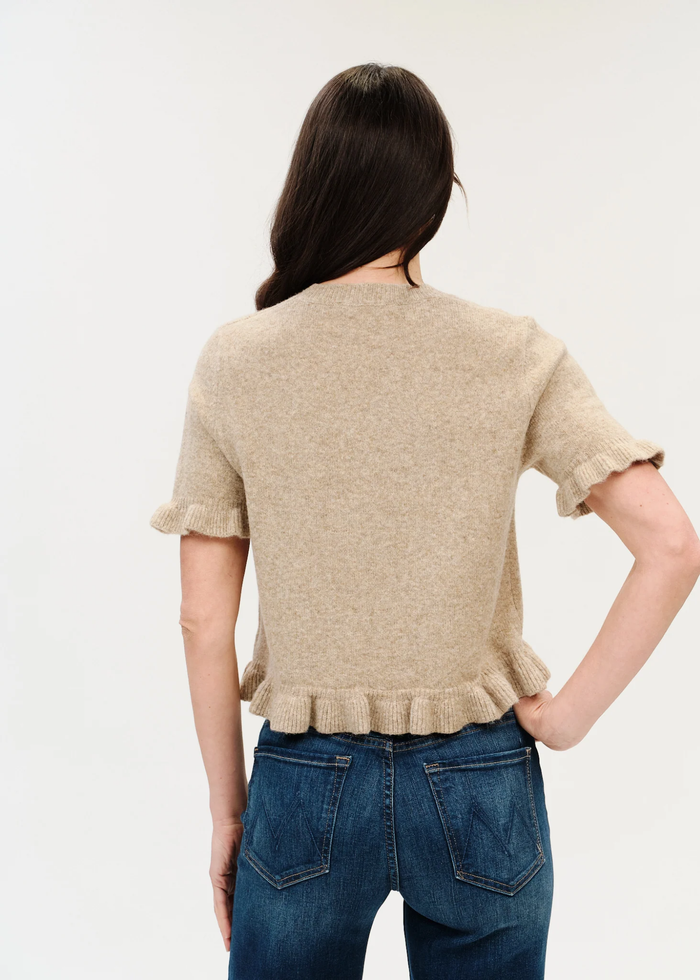 A woman viewed from the back wearing a beige short-sleeve knit top with ruffled edges and blue jeans, against a plain light background.