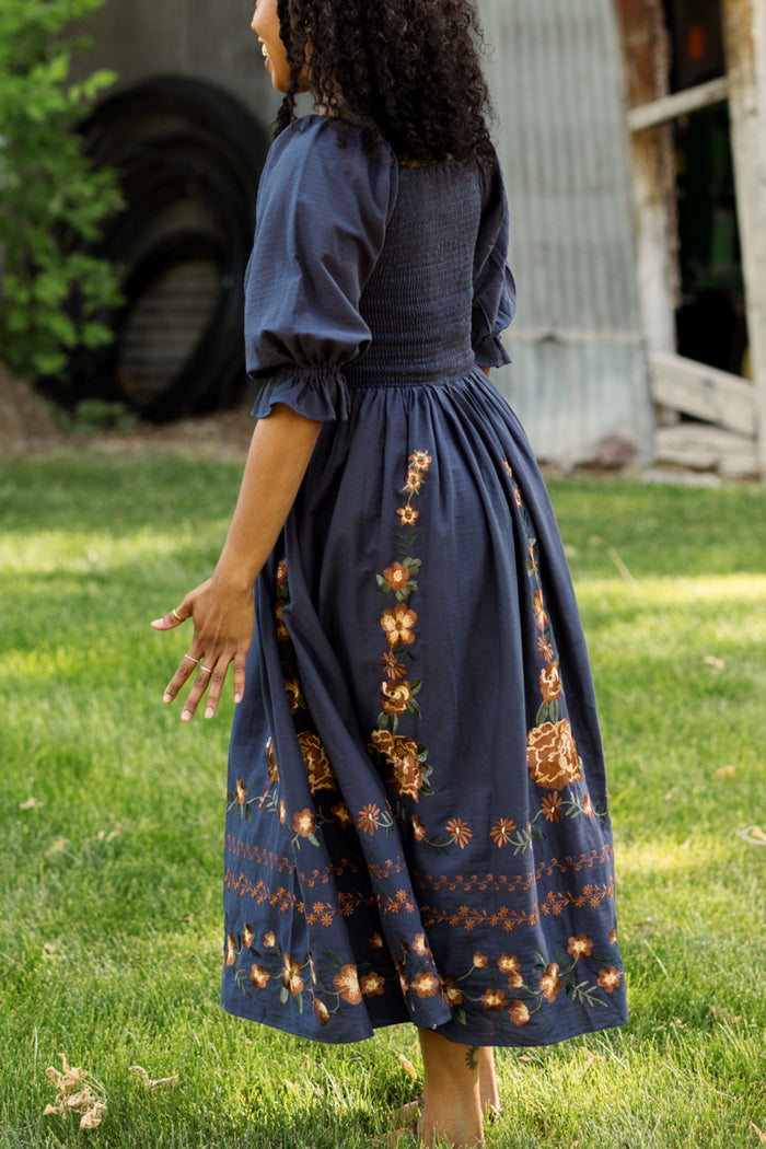 A woman with curly hair, barefoot on the grass and facing away from the camera, strolls near a corrugated metal shed and green trees. She wears the Ivy City Co Teagan Dress in Navy Embroidered Fall Floral, featuring a smocked bodice and vibrant floral patterns. The dress is maternity friendly, offering both comfort and style to her relaxed walk.