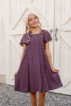A smiling young girl stands on gravel in front of a white wooden gate, holding out her Ivy City Co Teen Blakely Dress in Aubergine Ditsy Dot, which features a smocked bodice and flutter sleeves.
