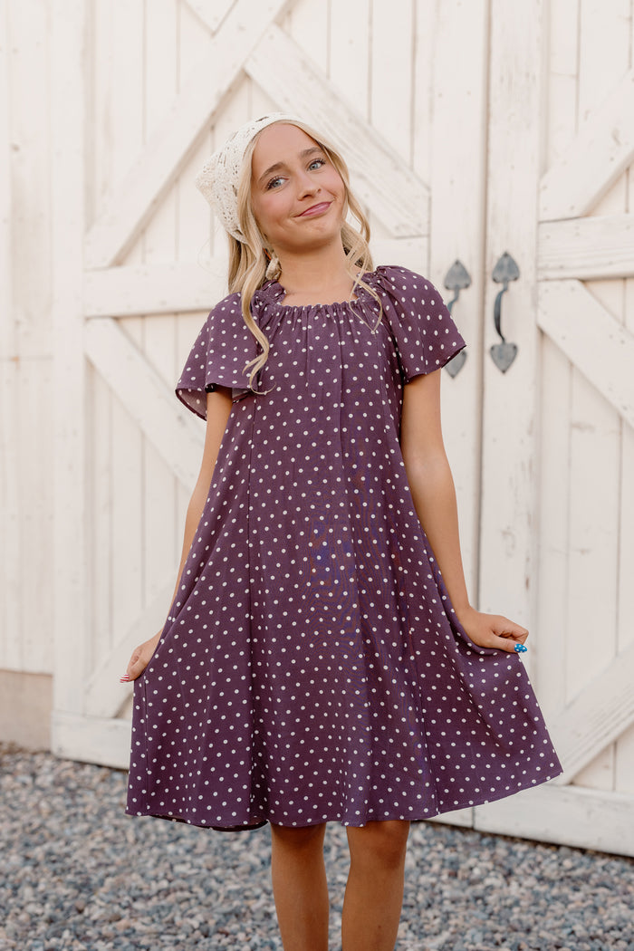 A smiling young girl stands on gravel in front of a white wooden gate, holding out her Ivy City Co Teen Blakely Dress in Aubergine Ditsy Dot, which features a smocked bodice and flutter sleeves.