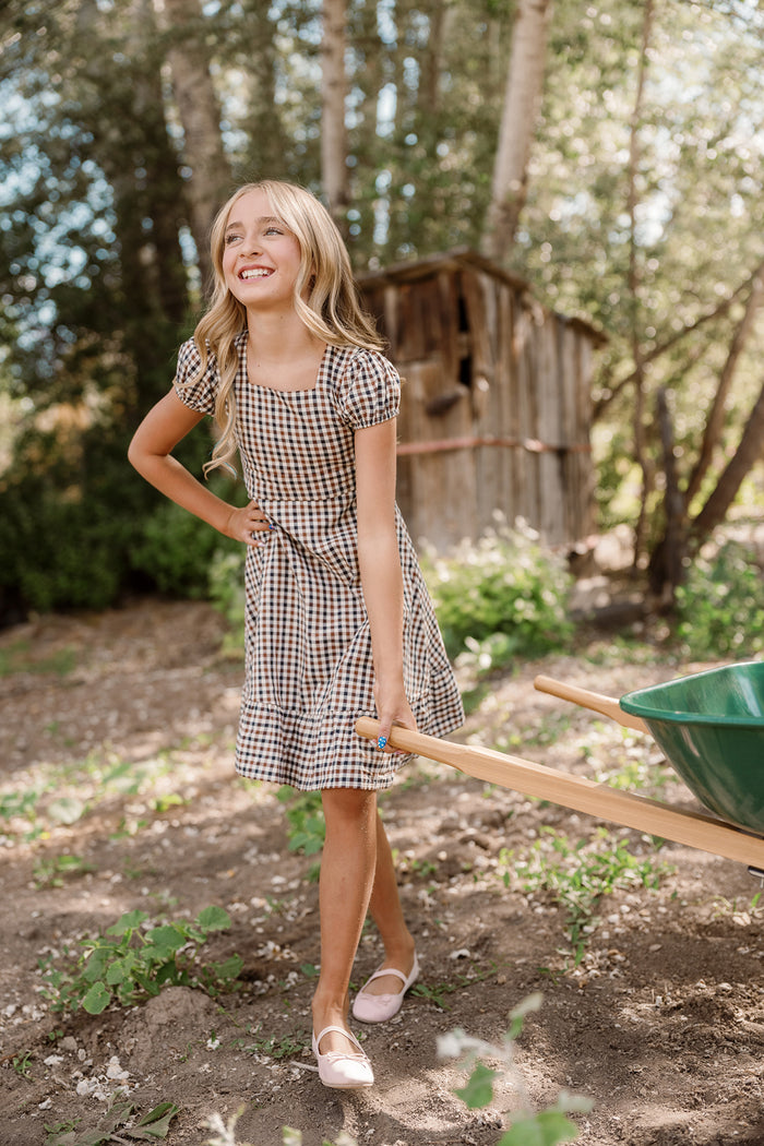 A smiling young girl stands outdoors on a sunny day, wearing the Ivy City Co Teen Connie Short Sleeve Dress in Chestnut Gingham. She holds a green wheelbarrow, surrounded by trees and with a small wooden shed in the background.