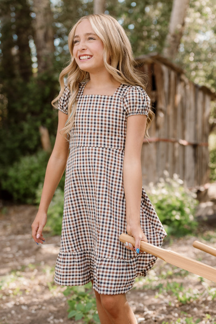 A young girl wearing the Ivy City Co Teen Connie Short Sleeve Dress in Chestnut Gingham smiles outdoors as she holds the handle of a wooden tool. She stands before a backdrop of trees and a rustic wooden shed, sunlight streaming through the leaves.