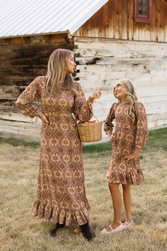 A woman and a girl, both wearing the Ivy City Co Teen Draper Dress in Golden Hour Tesserae, stand smiling and holding a basket outside a rustic wooden building on dry grass.