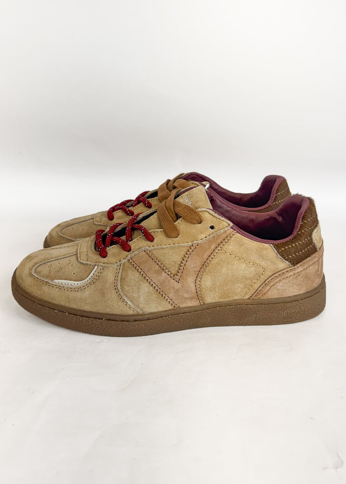 A pair of low-top sneakers in beige suede and tan leather, featuring brown laces with red tips, a brown sole, and a burgundy interior, shown against a plain white background.